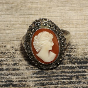 Op de afbeelding: Een zilveren ring met een cameo met het profiel van een vrouw. De cameo is gezet in een zilveren frame met een zwart-wit marmerpatroon.