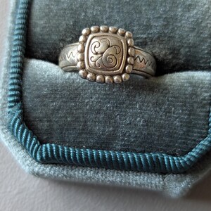 May include: Silver ring with a square, beaded centerpiece featuring an ornate swirl design. The ring is set in a teal velvet ring box. The band has a zig-zag pattern on each side of the square centerpiece.