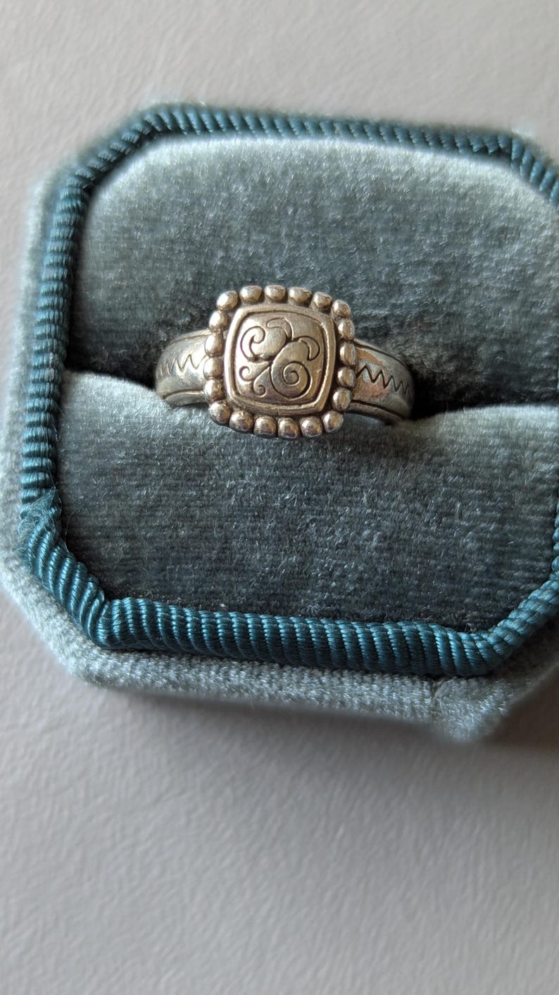 May include: Silver ring with a square, beaded centerpiece featuring an ornate swirl design. The ring is set in a teal velvet ring box. The band has a zig-zag pattern on each side of the square centerpiece.