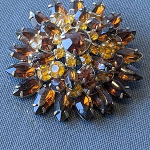 Könnte beinhalten: Eine große, runde Brosche aus goldfarbenem Metall, besetzt mit vielen braunen und bernsteinfarbenen Strasssteinen. Die Strasssteine sind in einem blumenartigen Muster angeordnet.