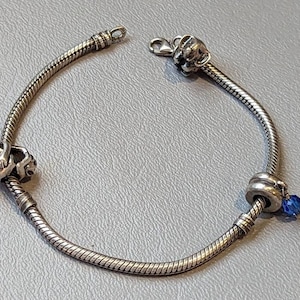 May include: A silver charm bracelet with three charms: a silver charm with a blue stone, a silver charm with a heart design, and a silver charm with a small animal design.