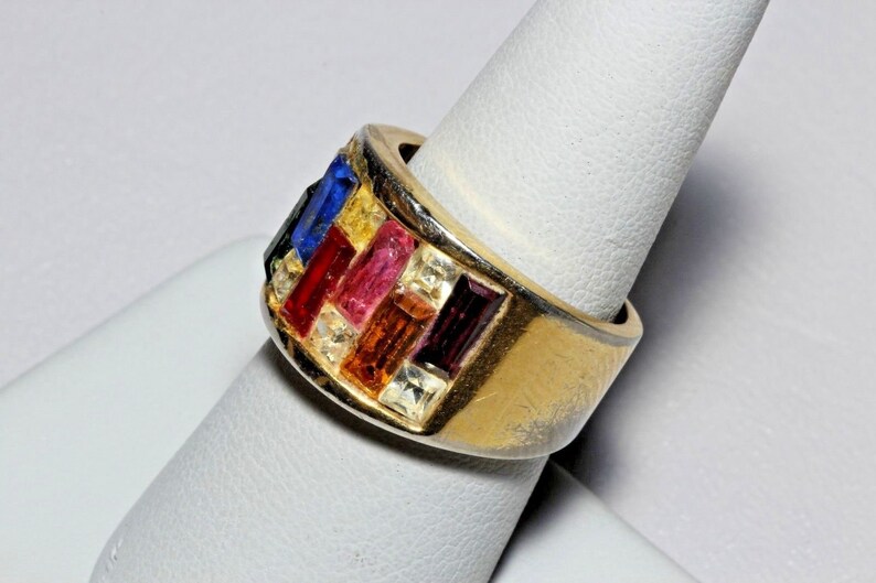 May include: A gold ring with a wide band set with colorful rectangular stones. The stones are arranged in a geometric pattern and include red, pink, yellow, blue, and clear stones.