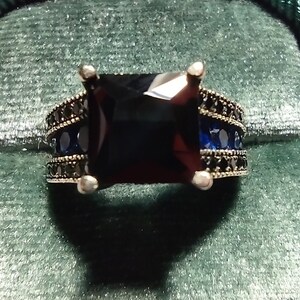 May include: A silver ring featuring a large, dark blue square-cut gemstone. The band is adorned with rows of smaller blue and black gemstones. The ring is displayed on a green velvet surface.