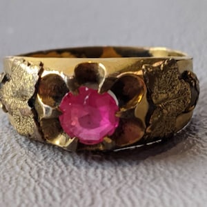May include: A gold ring with a pink gemstone set in a flower design. The ring is decorated with leaf-like details on the band.