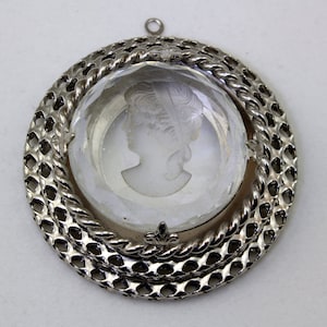 Puede incluir: Un colgante de plata con un camafeo de cristal transparente que representa el perfil de una mujer. El colgante está rodeado por un marco decorativo de plata.