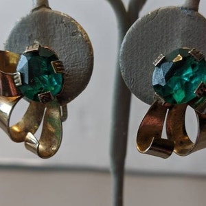 May include: A pair of gold tone clip-on earrings with green glass stones set in the center of each earring. The earrings are shaped like a bow.