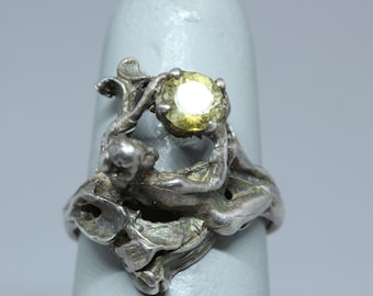 May include: A silver ring with a yellow gemstone set in a stylized design of two figures intertwined.