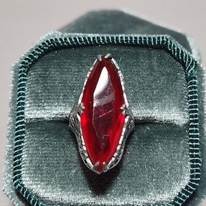 May include: A silver ring with a large, red, marquise-shaped gemstone set in a filigree design. The ring is displayed in a green velvet ring box.