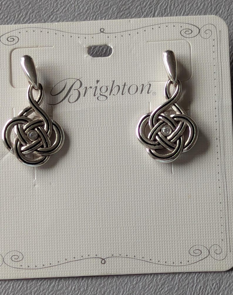 May include: A pair of silver-toned Brighton earrings. Each earring features a Celtic knot design with a small, clear crystal in the center. The earrings are displayed on a white card with the Brighton logo.