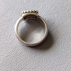 May include: A silver ring with a square-shaped top, adorned with small, round embellishments. The ring band has a simple, elegant design. The ring is set against a light gray textured surface.