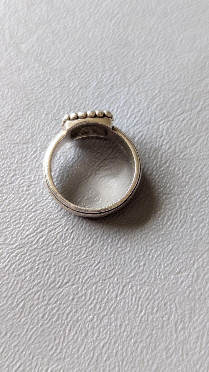 May include: A silver ring with a square-shaped top, adorned with small, round embellishments. The ring band has a simple, elegant design. The ring is set against a light gray textured surface.