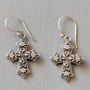 Può includere: Un paio di orecchini pendenti color argento con un ciondolo a forma di croce. Ogni croce presenta una superficie testurizzata con dettagli floreali e volute in rilievo. Gli orecchini sono fissati a semplici ganci curvi color argento.