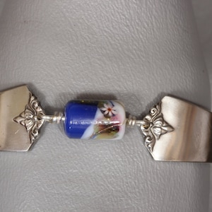 May include: A silver spoon bracelet with a blue and white glass bead. The bead has a floral design.