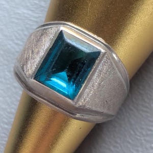 May include: Silver ring with a large, rectangular, bright blue gemstone. The ring has a textured, silver band and a classic, masculine design. The gemstone is set in a square bezel, creating a bold and eye-catching piece of jewelry.