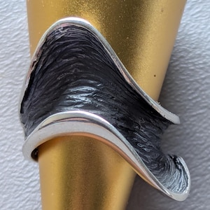 May include: A silver ring with a textured black center and a curved, wave-like design. The ring has a polished silver border and is set against a gold background. The ring's design is abstract and modern.