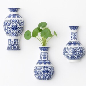 Puede incluir: Tres jarrones de cerámica blanca con diseños florales azules. Un jarrón contiene plantas verdes con hojas. Los jarrones tienen diferentes formas y tamaños, con diseños intrincados. Los jarrones están montados en una pared blanca.