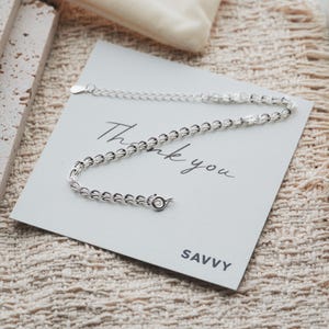 Pode incluir: Uma pulseira de corrente prateada com um design de elos delicados, apresentando um pequeno fecho de mola circular e uma corrente extensora ajustável. A pulseira é exibida num cartão branco com o texto Thank you e SAVVY impresso.