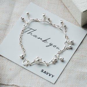 Puede incluir: Una pulsera de tono plateado con delicados dijes de metal en forma de lirios de los valles, dispuestos a lo largo de una cadena fina. La pulsera se presenta sobre una tarjeta cuadrada blanca con el texto Thank you y la marca SAVVY.
