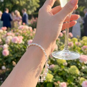 May include: A silver bracelet featuring a twisted metal band adorned with multiple small, dangling lily of the valley flower charms. The bracelet is worn on a wrist, with the metallic finish reflecting the surrounding light in an outdoor setting.
