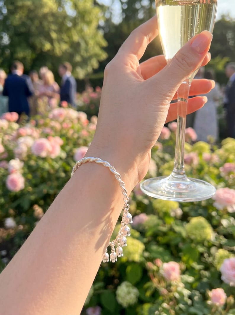 May include: A silver bracelet featuring a twisted metal band adorned with multiple small, dangling lily of the valley flower charms. The bracelet is worn on a wrist, with the metallic finish reflecting the surrounding light in an outdoor setting.