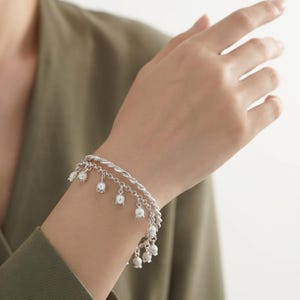 May include: A silver-toned double-strand bracelet featuring a twisted rope chain and a secondary chain adorned with multiple small, bell-shaped flower charms hanging at regular intervals along the length of the metallic jewelry piece.
