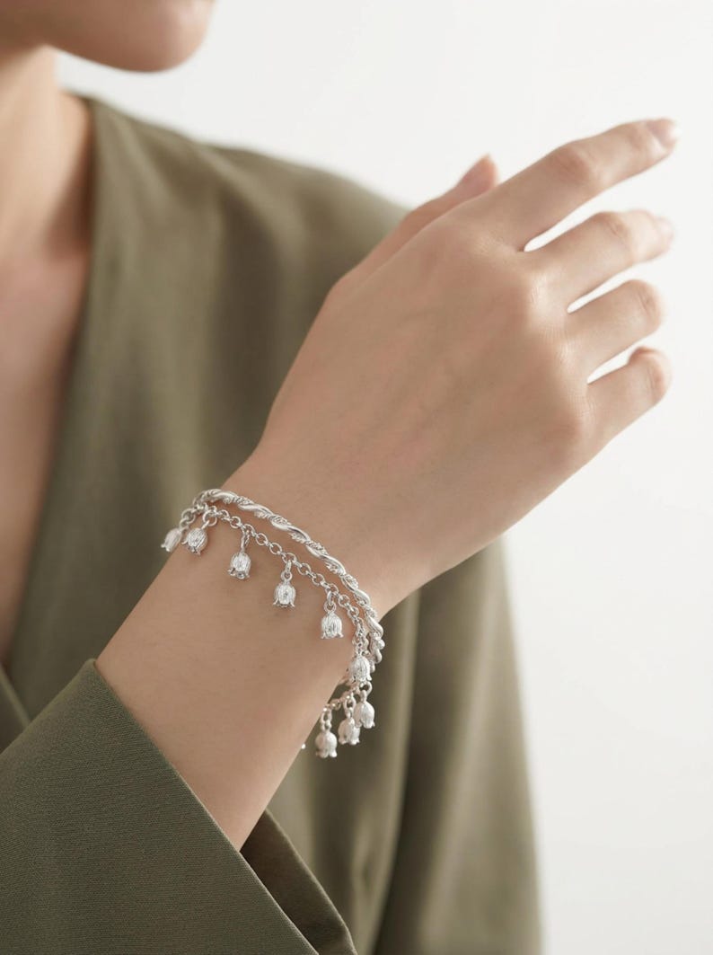 May include: A silver-toned double-strand bracelet featuring a twisted rope chain and a secondary chain adorned with multiple small, bell-shaped flower charms hanging at regular intervals along the length of the metallic jewelry piece.