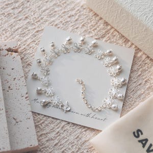 May include: A silver-toned bracelet featuring a repeating pattern of delicate open-work flowers and small bell-shaped charms. The bracelet is displayed on a square white card with the printed text Made by hand worn with heart.