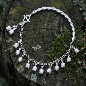 May include: A silver-toned bracelet featuring a series of small, bell-shaped flower charms hanging from a chain. The bracelet includes a twisted metal segment and a hook-style clasp, with multiple individual flower pendants dangling along the chain.