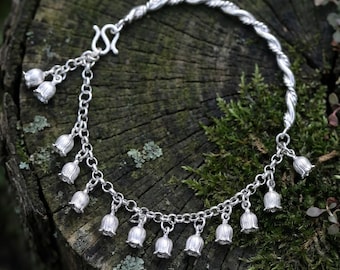 Peut inclure: Un bracelet de couleur argentée composé d'une chaîne ornée de petites breloques en forme de fleurs en cloche. Le bijou présente une section en métal torsadé, un fermoir à crochet et plusieurs pendentifs floraux suspendus le long du maillon.