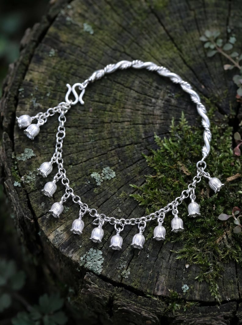 May include: A silver-toned bracelet featuring a series of small, bell-shaped flower charms hanging from a chain. The bracelet includes a twisted metal segment and a hook-style clasp, with multiple individual flower pendants dangling along the chain.