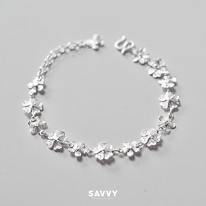 May include: A silver bracelet featuring a series of interconnected four-leaf clover charms with a textured, metallic finish. The piece includes a secure hook clasp and is marked with the text SAVVY at the bottom of the frame.