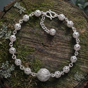 Puede incluir: Una pulsera de plata con una serie de esferas huecas de diseño intrincado, conectadas por delicados eslabones. La pulsera tiene un cierre decorativo y una cadena ajustable. Las esferas presentan patrones detallados.
