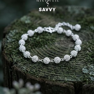 Può includere: Un braccialetto d'argento con perline sferiche dal design intricato. Il braccialetto è esposto su una superficie di legno muschiosa. Il testo "MISTVEIL x SAVVY" è visibile nella parte superiore dell'immagine.