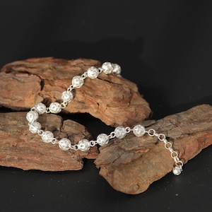 May include: A silver bracelet featuring a series of intricately designed, hollow spheres. The spheres are connected by small links, and the bracelet is displayed on a piece of textured wood against a dark background. The bracelet has a delicate, elegant appearance.