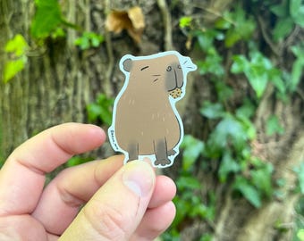 May include: A brown capybara sticker with a small cookie in its mouth. The sticker has a white border and is held in a hand against a background of green leaves and tree bark.
