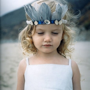 Puede incluir: Una niña joven con cabello rubio rizado que lleva un vestido blanco y una diadema de plumas plateadas y azules con pedrería.
