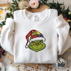 May include: White sweatshirt featuring a green cartoon character with an angry expression and a colorful Santa hat. The hat has red, green, yellow, and blue sections. The sweatshirt has a crew neck and long sleeves.