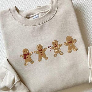 Puede incluir: Sudadera beige con cuatro hombres de jengibre bordados, con bufandas rojas y pajaritas. Uno de los hombres de jengibre sostiene un bastón de caramelo. Cuello redondo.