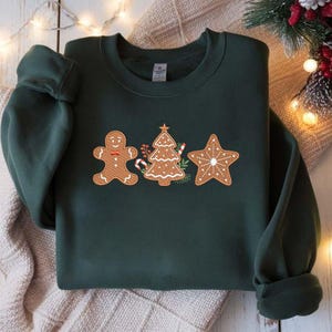 Op de afbeelding: Donkergroene sweatshirt met geborduurde peperkoekontwerpen. Het ontwerp omvat een peperkoekman, een kerstboom en een ster, allemaal in bruin- en wittinten. De sweatshirt wordt gepresenteerd op een neutrale ondergrond met kerstverlichting.