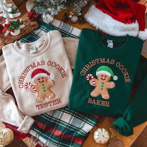 Könnte beinhalten: Zwei Sweatshirts, eines in Beige und eines in Grün, mit Weihnachtsstickerei. Jedes zeigt einen Lebkuchenmann mit Weihnachtsmütze und Zuckerstange, mit den Worten "Christmas Cookie" darüber. Das beige Sweatshirt trägt die Aufschrift "Tester" und das grüne "Baker".
