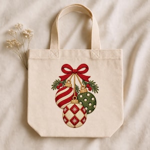 Peut inclure: Un sac fourre-tout en toile couleur cr&egrave;me orn&eacute; d'une broderie repr&eacute;sentant trois boules de No&euml;l suspendues, aux tons rouge, vert et blanc. Les boules sont nou&eacute;es par un ruban rouge et d&eacute;cor&eacute;es de branches de pin vertes et de baies rouges.