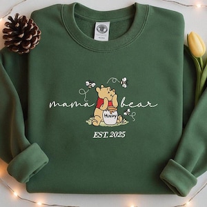 May include: A dark green sweatshirt featuring the embroidered phrase "mama bear" in white script. The design includes a cartoon bear, a honey pot, and bees. The text "EST. 2025" is below the bear.