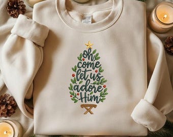 May include: Cream-colored sweatshirt with the text "Oh come let us adore Him" in a Christmas tree design. The design includes a gold star, green leaves, red berries, and a manger. The sweatshirt is surrounded by candles and pinecones.