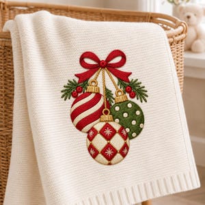 Peut inclure: Une couverture en tricot couleur cr&egrave;me orn&eacute;e d'une broderie repr&eacute;sentant trois boules de No&euml;l suspendues, aux tons rouge, blanc et vert. Les boules sont nou&eacute;es par un ruban rouge et d&eacute;cor&eacute;es de branches de pin vertes et de petites baies rouges.