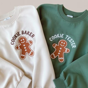 May include: Two crewneck sweatshirts, one cream and one green, with gingerbread man graphics. The cream sweatshirt says "COOKIE BAKER" and the green one says "COOKIE TESTER". The gingerbread men are brown with white outlines.