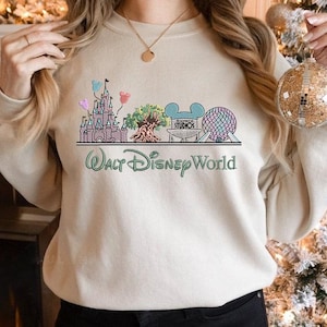 May include: A beige sweatshirt featuring a colorful graphic of Walt Disney World landmarks, including a castle, tree, and Spaceship Earth. The words "Walt Disney World" are printed in green script below the graphic. The sweatshirt has long sleeves.