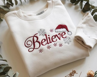 May include: Cream-colored sweatshirt with the word "Believe" embroidered in burgundy script, accompanied by snowflakes and a Santa hat. A matching sleeve is visible. The design is festive and suitable for the holiday season.