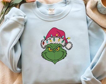 Op de afbeelding: Lichtblauw sweatshirt met een geborduurd Grinch-gezicht met een kerstmuts versierd met kerstverlichting. Het gezicht van de Grinch is groen met gele ogen en een ondeugende grijns. De hoed is rood met witte rand.