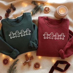 Könnte beinhalten: Zwei Rundhals-Sweatshirts, eines in Dunkelgrün und eines in Burgunderrot, jeweils mit einem weißen Lebkuchenmann-Design. Die Sweatshirts werden auf einer neutralen Oberfläche mit festlichen Dekorationen wie Tannenzapfen und getrockneten Orangenscheiben präsentiert.