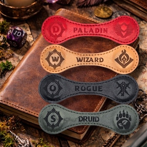 Pode incluir: Quatro fichas de classe de jogo em couro vermelho, bege, preto e verde. Cada ficha tem o formato de um haltere e apresenta um nome e símbolo de classe: Paladino, Mago, Ladino e Druida. As fichas estão em um livro encadernado em couro.